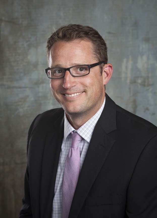 headshot of man in eyeglasses, black blazer, button up and lavender tie