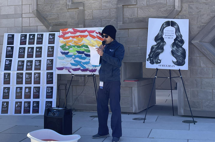 person speaking during rally in front of poster reading I Stand with the Women of Iran