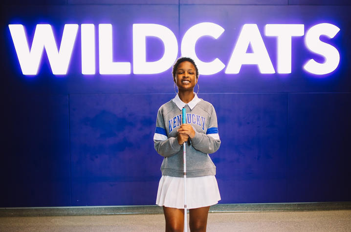 Girl in grey sweater standing in front of blue wall with "WILDCATS" on the wall.