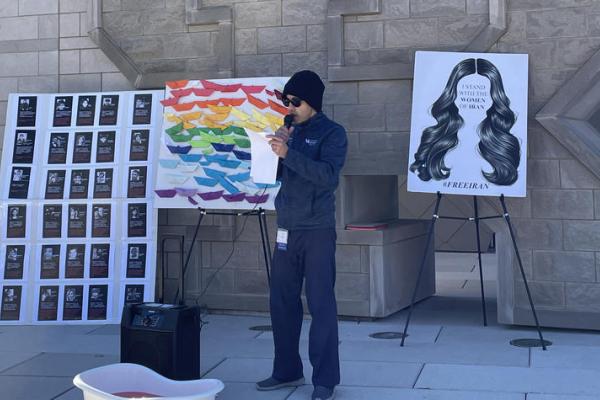 person speaking during rally in front of poster reading I Stand with the Women of Iran