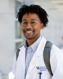 image of Black student wearing glasses, white coat, blue button up and a backpack